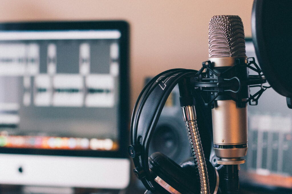 A studio microphone with headphones is set up in front of a computer monitor displaying audio editing software.